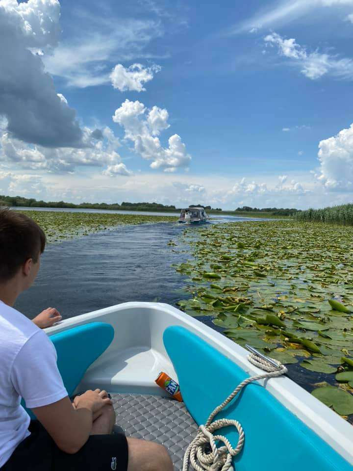 Nearchus Nautica Excursii in Delta O persoană într-un tricou alb și pantaloni scurți negri stă pe o barcă, înconjurată de apă și nufăr sub un cer parțial înnorat. În depărtare se vede o altă barcă. Fața bărcii are o frânghie încolăcită și un pachet de gustări este în fața persoanei, care se bucură de plimbari cu barca în Delta Dunarii cu nearchus Nautictour.