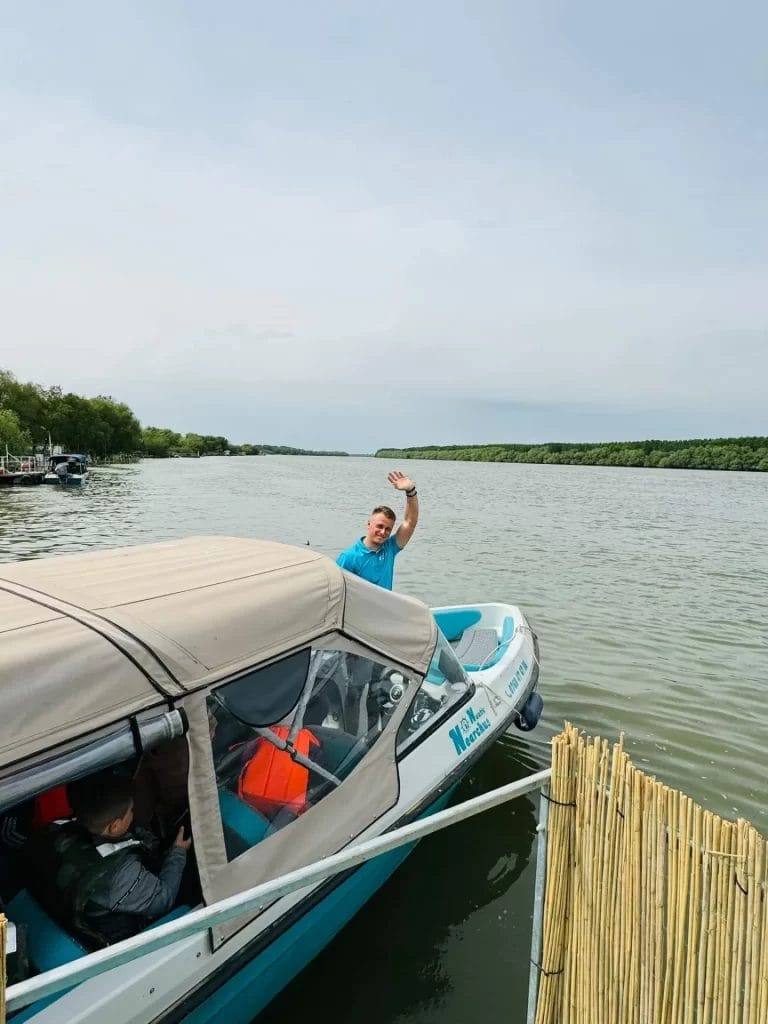 Nearchus Nautica Excursii in Delta O persoană face cu mâna de pe o barcă cu motor mică, albă și albastră, acostată de un gard natural, din lemn, pe un râu calm, cu maluri verzi, mărginite de copaci. Barca are un baldachin bej. Cerul este înnorat, iar în stânga se vede o altă barcă mai jos pe râu, parte din traseu croaziere din Delta Dunarii.