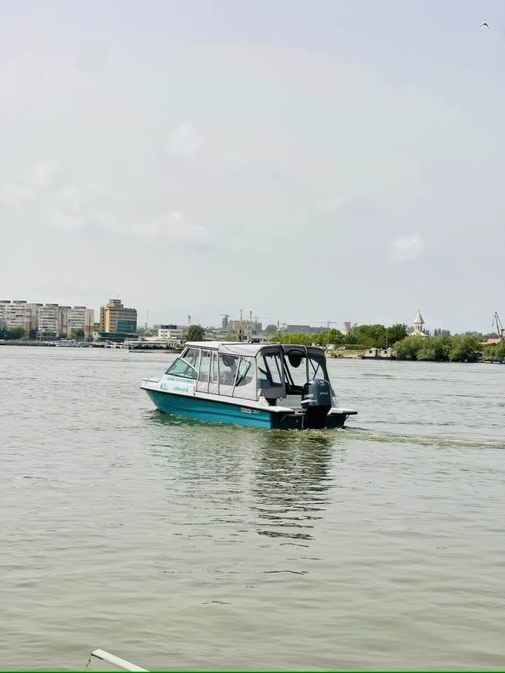 Nearchus Nautica Excursii in Delta O barcă cu motor mică, albastră și albă, probabil o barca metalica de la Nearchus NauticTour Mahmudia, navighează pe ape calme lângă țărmul unui oraș. Clădiri urbane, copaci verzi și o biserică albă îndepărtată cu o cupolă sunt vizibile în fundal sub un cer parțial înnorat. Scena este senină, cu ondulații blânde în spatele bărcii.