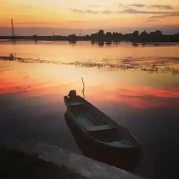 Nearchus Nautica Excursii in Delta O barcă de lemn este ancorată pe malul unui lac liniştit în timpul apusului. Cerul afișează nuanțe de portocaliu, roz și galben, reflectându-se frumos pe apă. Copacii și bărcile cu pânze sunt vizibile în depărtare pe fundalul senin.