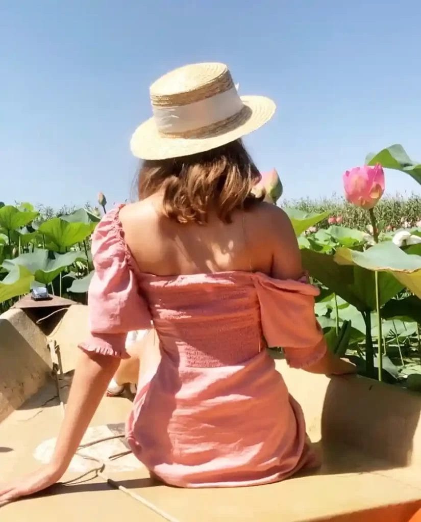 Nearchus Nautica Excursii in Delta O femeie care poartă o pălărie de paie și o rochie roz deschis fără umeri stă pe o barcă înconjurată de frunze verzi mari și flori de lotus roz sub un cer albastru senin. Ea este cu fața departe de cameră, bucurându-se de peisajul natural senin.