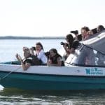 Nearchus Nautica Excursii in Delta Un grup de turisti in vacanta in Delta Dunarii pe o mica barca cu motor alb-alb, numita "Nautic Researchus", fac fotografii cu ceva din stanga. Ei țin în mână camere cu lentile lungi, concentrate atent. Apa este calmă, iar cerul este senin.