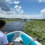 Nearchus Nautica Excursii in Delta O persoană într-un tricou alb și pantaloni scurți negri stă pe o barcă, înconjurată de apă și nufăr sub un cer parțial înnorat. În depărtare se vede o altă barcă. Fața bărcii are o frânghie încolăcită și un pachet de gustări este în fața persoanei, care se bucură de plimbari cu barca în Delta Dunarii cu nearchus Nautictour.
