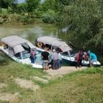 Un grup de oameni stă la marginea unui râu lângă două bărci cu motor care sunt acostate una lângă alta, gata de excursii în Delta Dunării. Unii indivizi se urcă în bărci, în timp ce alții așteaptă pe malul înierbat. Bărcile au copertine, iar râul este înconjurat de verdeață luxuriantă, inclusiv diverse planta din Delta Dunarii. Vremea pare a fi însorită și plăcută.