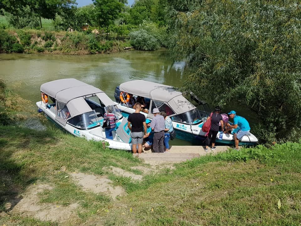 Un grup de oameni stă la marginea unui râu lângă două bărci cu motor care sunt acostate una lângă alta, gata de excursii în Delta Dunării. Unii indivizi se urcă în bărci, în timp ce alții așteaptă pe malul înierbat. Bărcile au copertine, iar râul este înconjurat de verdeață luxuriantă, inclusiv diverse planta din Delta Dunarii. Vremea pare a fi însorită și plăcută.