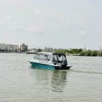 Nearchus Nautica Excursii in Delta O barcă cu motor mică, albastră și albă, probabil o barca metalica de la Nearchus NauticTour Mahmudia, navighează pe ape calme lângă țărmul unui oraș. Clădiri urbane, copaci verzi și o biserică albă îndepărtată cu o cupolă sunt vizibile în fundal sub un cer parțial înnorat. Scena este senină, cu ondulații blânde în spatele bărcii.