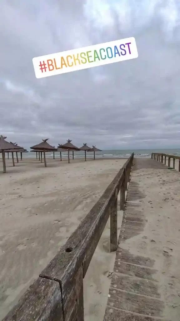 Nearchus Nautica Excursii in Delta Promenadă de lemn care duce la o plajă de nisip cu adăposturi în formă de umbrelă. Cerul este înnorat, sugerând vreme înnorată. Marea este pe fundal. Un hashtag în text de culoarea curcubeului scrie „#BLACKSEACOAST” în partea de sus a imaginii.