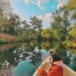 Nearchus Nautica Excursii in Delta O persoană într-o jachetă portocalie stă la prova unei bărci mici, ținând în mână o cameră, navigând într-un corp de apă calm, înconjurat de copaci luxurianți. O casă în copac este vizibilă printre copacii din stânga. Cerul este parțial înnorat, reflectându-se frumos pe apa liniștită de dedesubt.