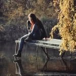 Nearchus Nautica Excursii in Delta O femeie cu păr lung și negru, purtând un pulover închis la culoare și blugi, stă pe marginea unui doc de lemn deasupra unui corp de apă nemișcat. Ea se uită în jos și atinge apa cu mâna. Frunzișul de toamnă cu frunze aurii atârnă în dreapta, reflectându-se ușor pe suprafața apei.